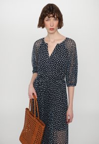 Mørkeblå, gjennomsiktig dagkjole med blomsterdesign, V-hals, puffermer, og rynket midje; accessorert med en vevd brun veske.