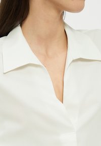 Γυναίκα που φοράει λευκό πουκάμισο με γιακά και ανοιχτό ντεκολτέ, ορατό από τον λαιμό έως το επάνω μέρος του στήθους, σε απλό φόντο.