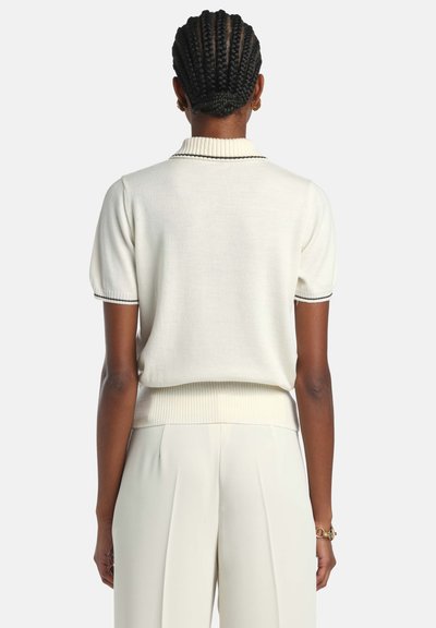 Kremowa krótka dzianinowa koszulka polo z prążkowanym kołnierzykiem i dołem, z czarnymi liniami akcentującymi. Gładka tekstura i luźny krój, zestawiona z jasnymi spodniami.