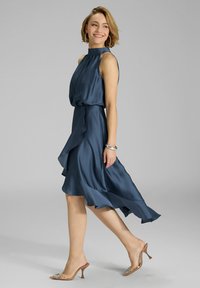 Robe en satin bleu marine avec un col haut, un ourlet volanté et une silhouette fluide, associée à des sandales à talons transparents et des bijoux en argent.