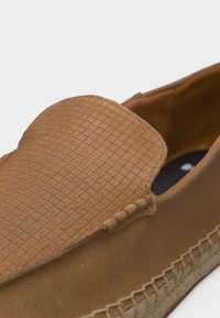 Κοντινή λήψη του καφέ δερμάτινου μοκασίνι με επάνω μέρος από σουέτ και πλεκτή λεπτομέρεια κατά μήκος της άκρης της σόλας.