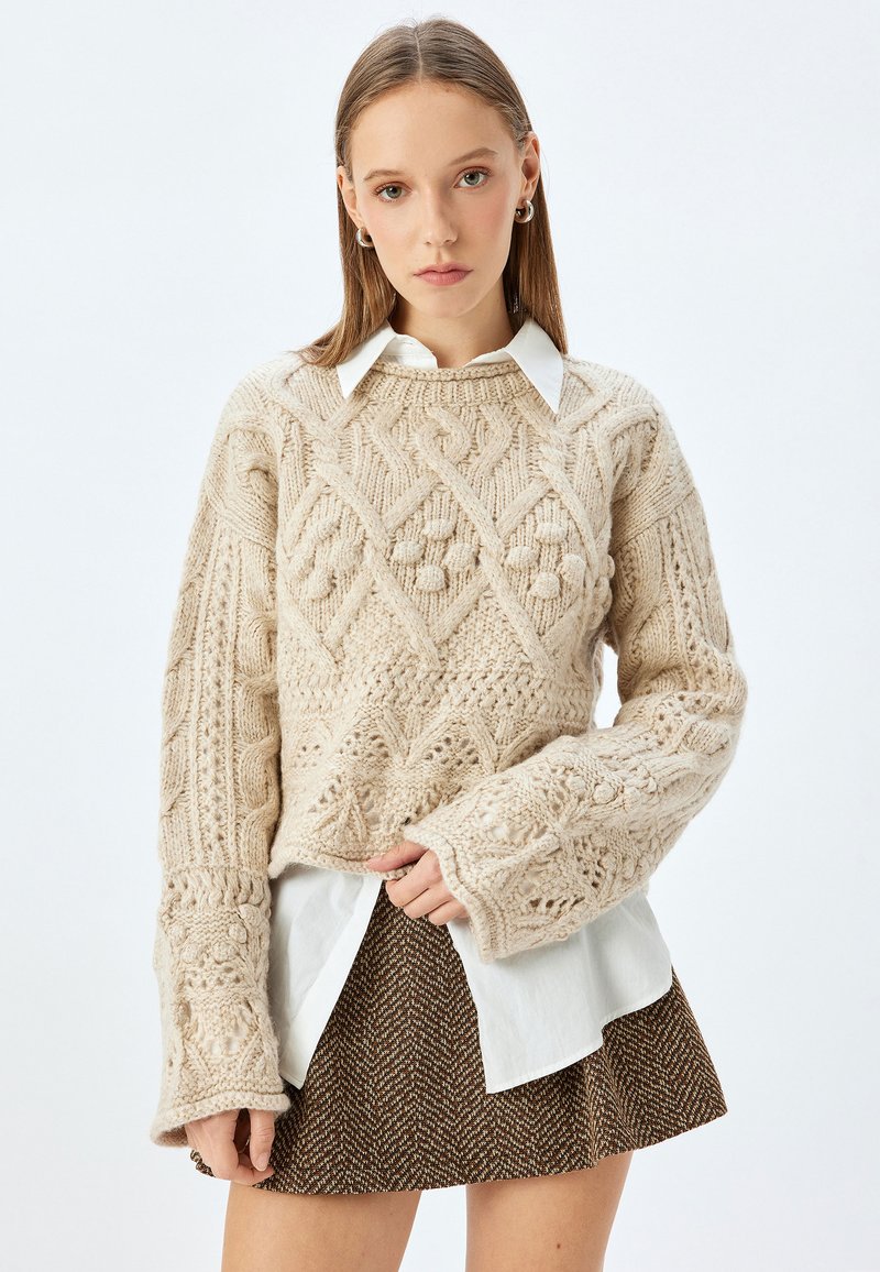 Maglione beige a maglia con un motivo testurizzato, maniche larghe, indossato sopra una camicia bianca e abbinato a una gonna marrone a motivo.