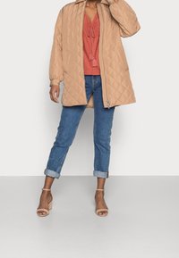 Beige tikkusu takki, jossa on kaulus, yhdistettynä ruosteenväriseen yläosaan. Siniset käärettävät farkut ja beige korkeat sandaalit täydentävät asua.