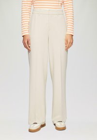 Persoon gekleed in crèmekleurige wijde broek, witte en bruine sneakers, en een langemouwen oranje en witte gestreepte top, staande tegen een effen achtergrond.