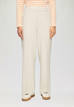 Personne portant un pantalon large crème, des baskets blanches et marron, et un haut à manches longues rayé orange et blanc, se tenant contre un fond uni.