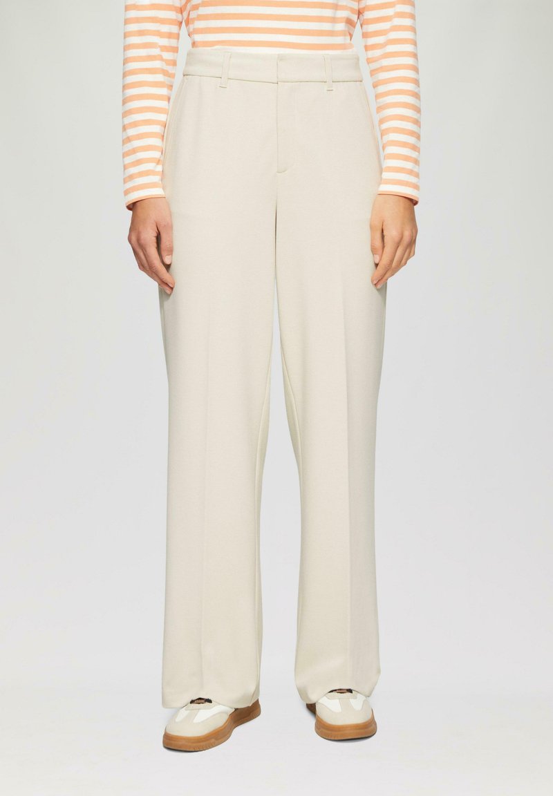 Persoon gekleed in crèmekleurige wijde broek, witte en bruine sneakers, en een langemouwen oranje en witte gestreepte top, staande tegen een effen achtergrond.