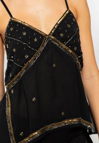 Top negro sin mangas con cuello en V, adornado con lentejuelas doradas y plateadas y patrones de estrellas, con una tela fluida y texturizada.