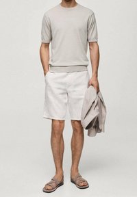 T-shirt à manches courtes en maille gris clair, short en lin beige et sandales grises. Le mannequin tient une veste assortie gris clair. Design simple et couleurs neutres.