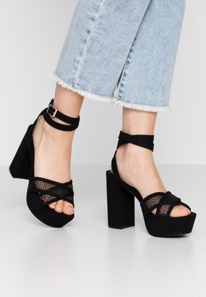 Pieds portant des sandales à plateforme noires à talons hauts avec des brides à la cheville, associées à un jean court bleu clair à ourlet effiloché sur un fond clair.