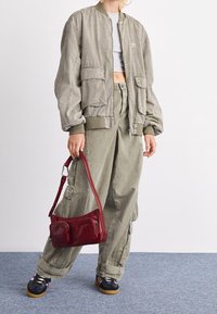 Olive green bomber jacket with two front pockets, paired with matching baggy cargo pants. Holds a small red leather handbag with zippers.