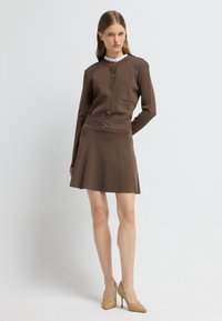Bruine trui-jurk met een aansluitend bovenstuk en een uitlopende rok, met gouden knopen en een taille-accent. Gdraagd met beige hoge hakken.