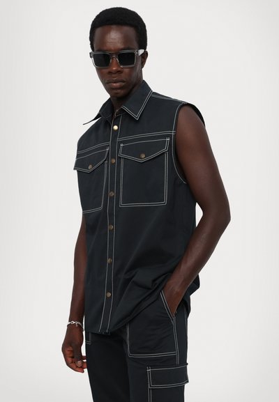 Black sleeveless shirt with front pockets, contrasting white stitching, and metal buttons. Worn with matching pants and sunglasses.