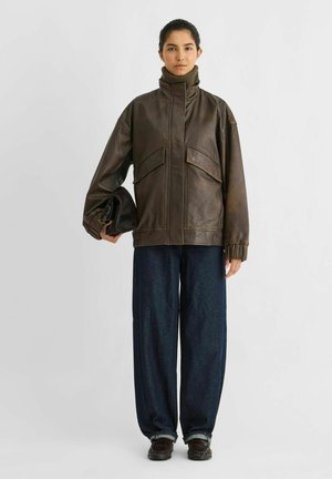 Le modèle se tient devant un fond de studio uni, portant une veste en cuir marron, un jean foncé à jambes larges et des chaussures, et tenant un sac noir.