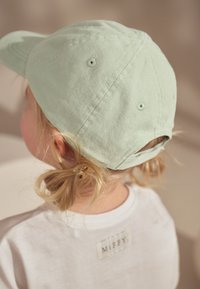 Cappellino di cotone verde menta con visiera curva e fori di ventilazione. Indossato al contrario, lasciando intravedere capelli biondo chiaro raccolti in due piccole code.