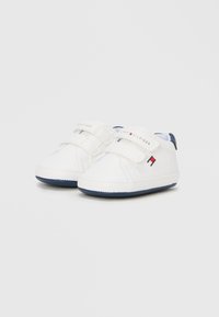 Λευκά παπούτσια για νήπια με δύο ιμάντες βέλκρο, ναυτικό μπλε λεπτομέρειες στη φτέρνα και ελαστική σόλα από καουτσούκ. Το λογότυπο εμφανίζεται στους ιμάντες. Λείο συνθετικό υλικό.