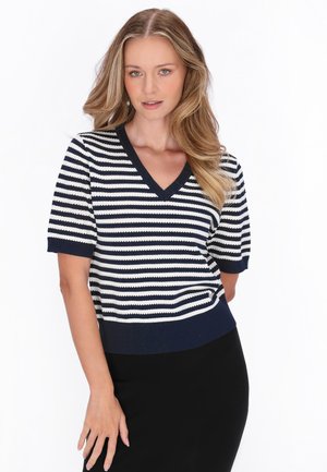 Frau mit langen blonden Haaren, die ein dunkelblau-weiß gestreiftes V-Ausschnitt-Kurzarmoberteil und einen schwarzen Rock vor weißem Hintergrund trägt.