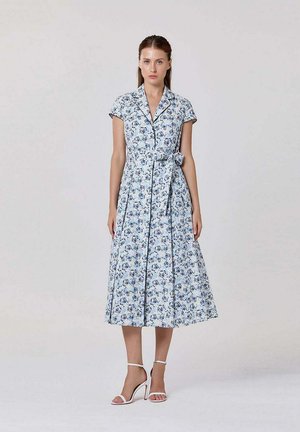 Femme debout portant une robe mi-longue à manches courtes avec col, motif floral bleu et ceinture nouée à la taille, associée à des sandales blanches à talons hauts.