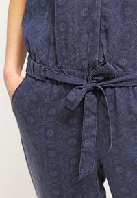 Primer plano de una persona que lleva pantalones oscuros de rayas azul con un cinturón de tela a juego atado en forma de lazo y las manos en los bolsillos.