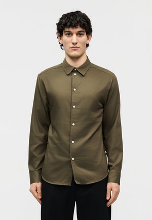 Noor mees lühikeste lokkis juustega, kes kannab oliivrohelise värvi pikakäist nööpidega särki ja musti pükse, seisab otsevaates ilma taustata.