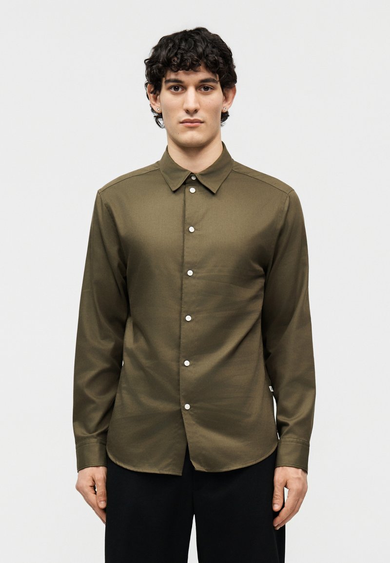 Νεαρός άνδρας με κοντά σγουρά μαλλιά, φορώντας μια πουκάμισο μακρυμάνικο ελιάς με κουμπιά και μαύρο παντελόνι, που κοιτάει μπροστά μπροστά σε απλό φόντο.