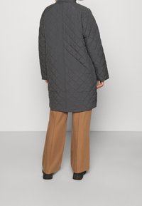 Lungo cappotto grigio imbottito con colletto alto, motivo a rombi e cerniere laterali. Indossato su ampi pantaloni beige, con un design dritto sul retro.