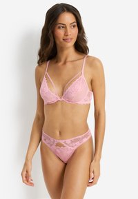 Γυναίκα με μακριά καστανά μαλλιά φορώντας ροζ δαντελένιο σουτιέν και αντίστοιχο δαντελένιο εσώρουχο, στέκεται μπροστά σε λευκό φόντο.