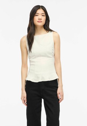 Vrouw met lang donker haar, die een mouwloos wit aansluitend topje en een zwarte broek draagt, staand tegen een effen witte achtergrond.