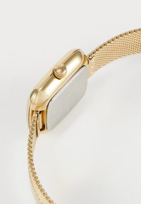 Montre avec bracelet en maille dorée, boîtier rectangulaire, finition texturée dorée et couronne proéminente. Le dos présente des détails gravés.
