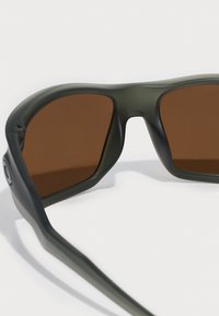 Ochelari de soare cu un cadru mat de culoare verde măsliniu și lentile cu tentă maronie. Design plat cu margini rotunjite. Vedere laterală care arată o formă aerodinamică.