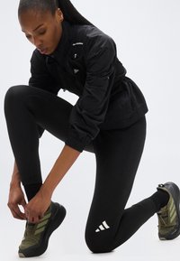 Γυναίκα με μαύρα αθλητικά ρούχα δένει τα ελιάς πράσινου χρώματος παπούτσια τρεξίματος, γονατίζοντας σε ένα γόνατο μπροστά σε απλό λευκό φόντο.