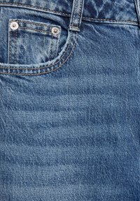 Gros plan sur un tissu en denim bleu avec une petite poche à monnaie munie de rivets métalliques et des coutures visibles sur un jean.