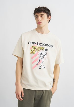 Joven con cabello rizado que lleva una camiseta crema de New Balance con un gráfico de un jugador de fútbol y el texto "Equipo Atlético Profesional".