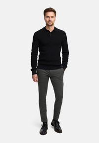 Schwarzes Langarm-Polohemd mit gerippter Textur, kombiniert mit schmal geschnittenen grauen Hosen und schwarzen Lederschuhen. Das Modell steht mit den Händen an den Seiten.