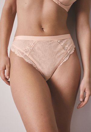 Naine kannab virsikuvärvilist pitsilist kõrge vöökohaga pesu ja sobivat rinnahoidjat, seistes neutraalse tausta ees.