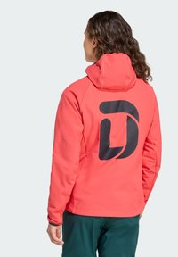 Koraalrode softshell jas met capuchon; heeft een groot zwart logo op de rug; gladde textuur, lange mouwen en een ontspannen pasvorm.