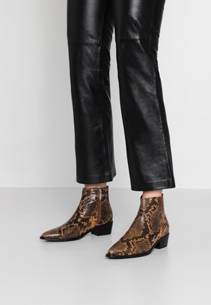 Knöchelhohe Stiefel aus Leder mit braun-schwarzem Schlangenhautmuster, getragen zu schwarzen, gekürzten Lederhosen.