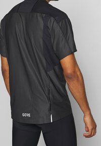 Černé sportovní tričko s krátkým rukávem, které má síťované a hladké panely, s zipem na zádech a logem na spodním lemu.