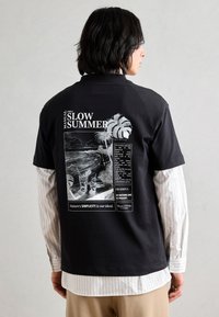 Black t-shirt with a graphic print on the back featuring tropical imagery and text. Short sleeves, relaxed fit, and cotton material.
