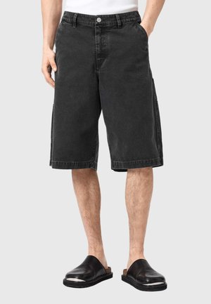 Czarne dżinsowe spodenki cargo o prostym kroju, z bocznymi kieszeniami i zapięciem na guzik. Noszone z czarnymi sandałami na płaskiej podeszwie, prezentowane na szarym tle.