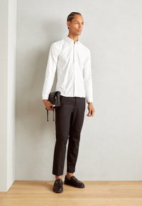Camisa branca de botão com colarinho, calças pretas slim-fit e sapatos pretos sem atacadores. Segurando uma pequena bolsa preta. Texturas suaves em todo o conjunto.