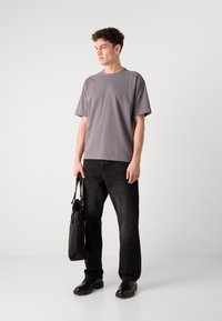 T-shirt gris à manches courtes associé à un jean noir, tenant un sac noir. Le tissu a l'air lisse et la coupe est décontractée. Les chaussures sont noires.