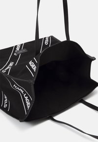 Czarna torba na ramię wykonana z wytrzymałego materiału, z białymi wzorami logo, długimi płaskimi uchwytami i niepodszewkowanym wnętrzem. Otwarte wzornictwo na górze.