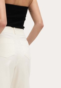 Vrouw in hoge witte broeken en een zwart mouwloos geribbeld topje, gezien van achteren tegen een effen achtergrond.