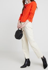 Oranssi vanutettu vetoketjuneule, kermaiset suorat housut, musta pyöreä käsilaukku ja mustat teräväkärkiset nilkkasaappaat. Yksinkertainen muotoilu ilman kuvioita.