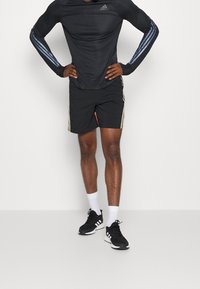 Camisa atlética de manga larga negra con franjas azules y logo, combinada con pantalones cortos negros con acentos dorados y un bolsillo con cremallera. Zapatillas deportivas negras.