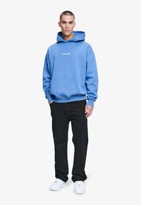 Sudadera de algodón azul con un bolsillo frontal, que presenta un logo blanco de "PEGADOR". Combinada con pantalones negros y zapatillas blancas. Textura suave.