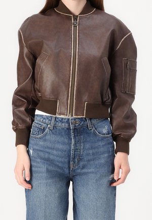 Vrouw draagt een cropped donkerbruine leren bomberjack met een ritssluiting aan de voorkant en blauwe high-waisted jeans met lichte slijtageplekken.