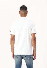 Weißes Poloshirt mit kurzen Ärmeln, Kragen und Logo auf dem linken Ärmel, kombiniert mit hellblauen Jeans. Glatte Textur, lässiges Design.