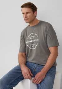 Grijze katoenen t-shirt met korte mouwen, voorzien van witte tekst in een cirkelontwerp, gecombineerd met vervaagde blauwe denim jeans. Minimalistische esthetiek.