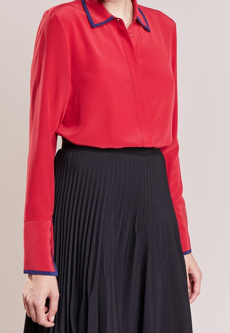 Personne portant une chemise rouge à manches longues avec un bord bleu sur le col et les poignets, rentrée dans une jupe noire plissée taille haute.